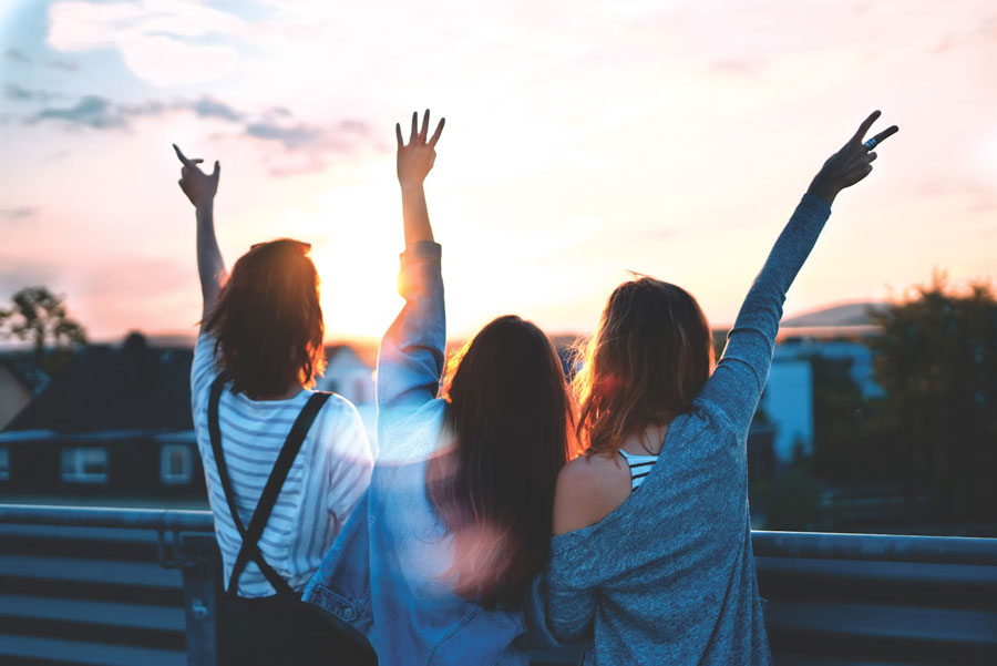 Three women stand on a rooftop at sunset, with arms raised and peace signs. 