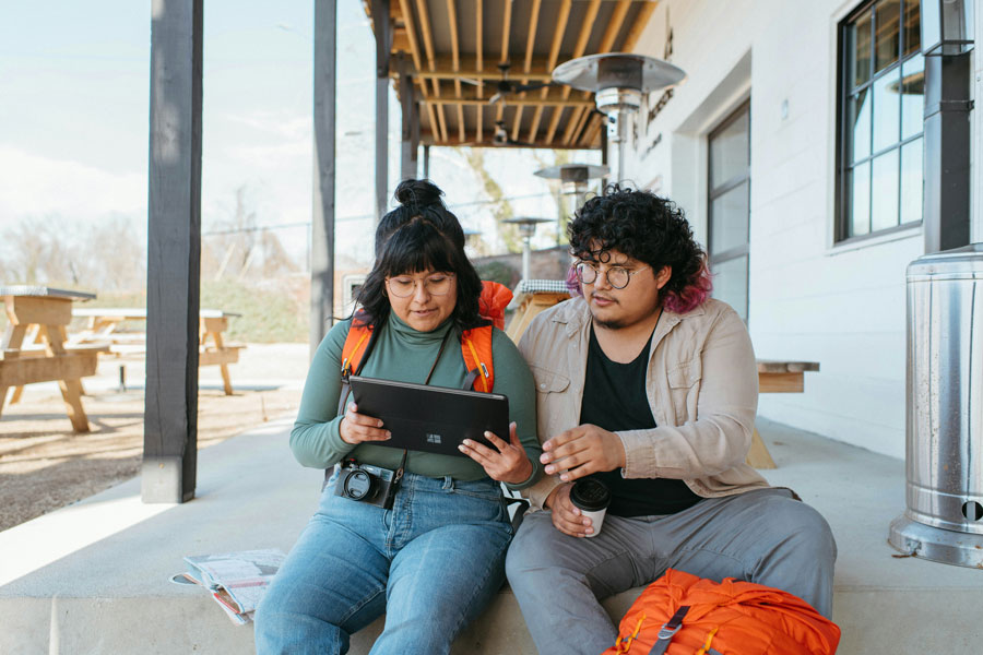Two people sit outside on a porch, engaged with a tablet. One wears an orange backpack, and the other holds a drink. The scene is casual and relaxed.